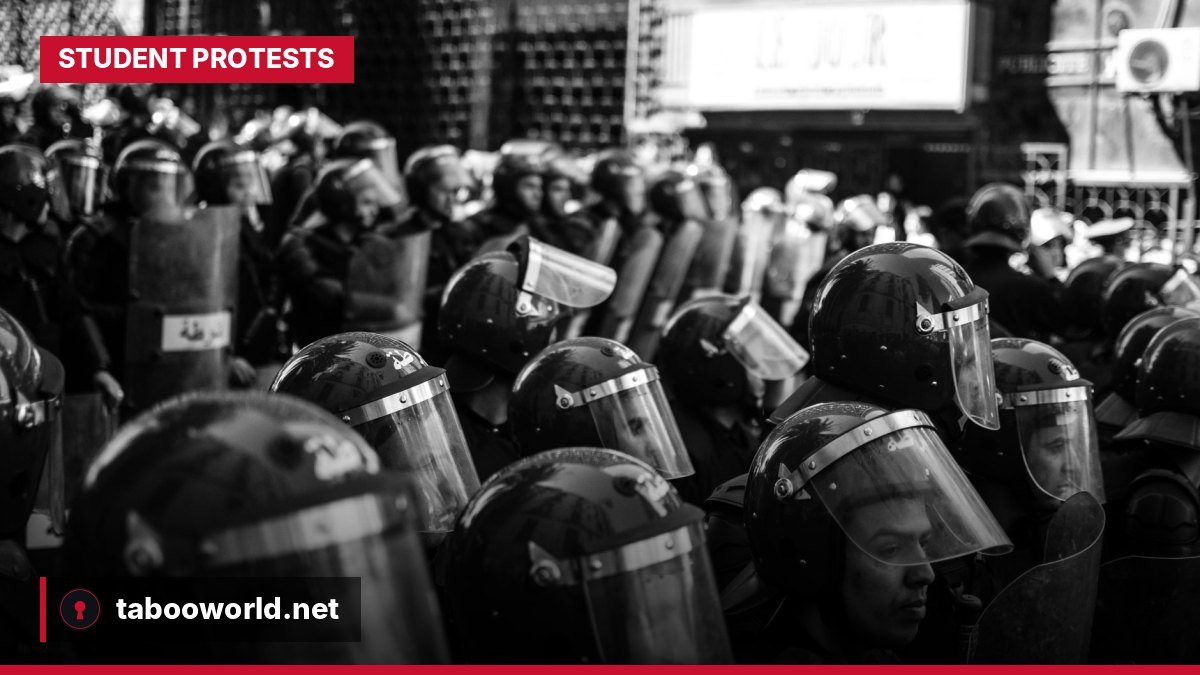 Riot police with shields facing a group of young, masked protesters in a city street in France.